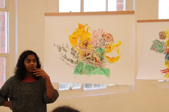 A person of colour with dark hair in a grey sweater stands to the left of the image in the bottom corner, gesturing their hand toward their mouth in conversation. They look to their left implying the presence of another figure. Behind them are 2 suspended artworks hanging using wire, from wooden beams. They are paper works with large bursts of colour, very intricate mark makings.