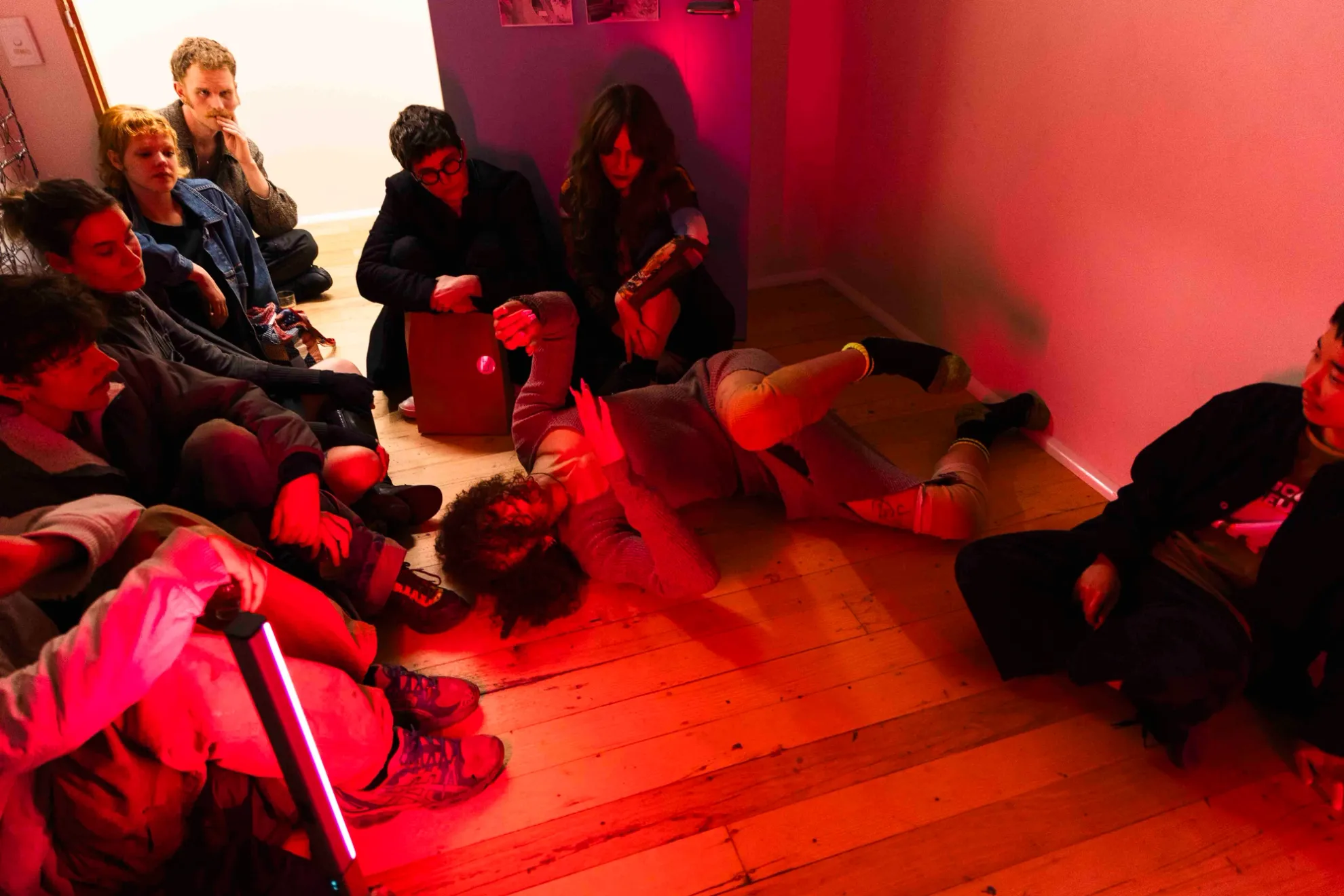 A group of people watching Andrea Illes perform in a small room bathed in red light. Andrea is rolling around on the floor performing