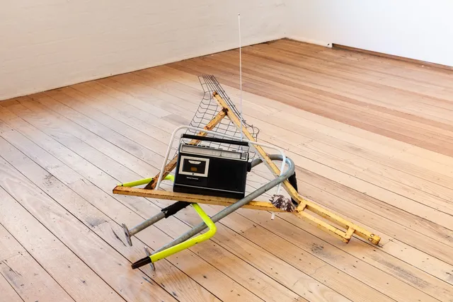 Sculptural installation piece comprised of metal poles, a brown shell, wire frame and a radio sits on the warm floorboards of the gallery.