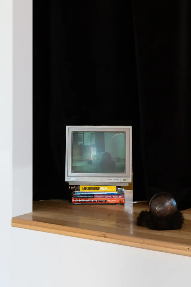 Installation view of "Big Pharmakon". The image shows a small analog television sitting on a pile of books on a wooden counter. The screen displays a live camera feed of the West Space Gallery. In front of the television is a small ceramic ball resting on possum fur. A black curtain hangs behind the counter.