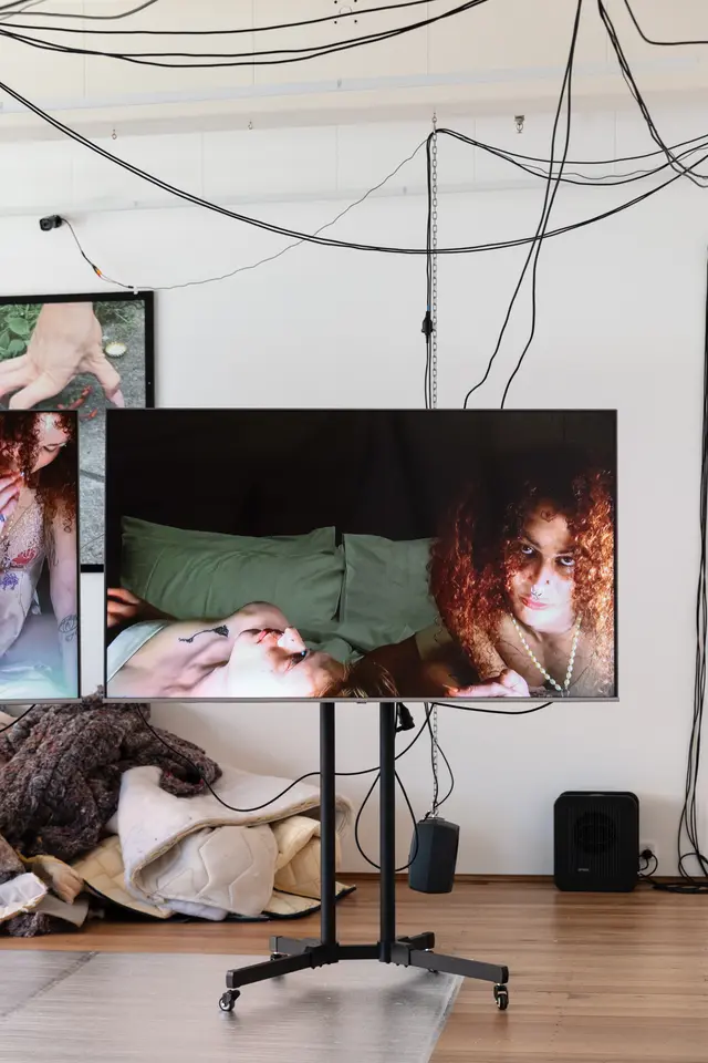 A woman and another person is displayed on a screen. A pile of textiles from the mattress and an image can be seen in the background.
