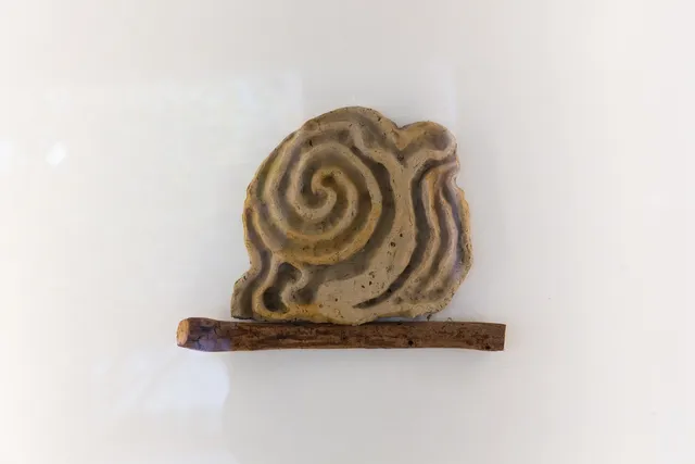 A sculpted slab of clay that resembles a walking labrynth sits upon a stick that is installed to wall of the West Space Window gallery.