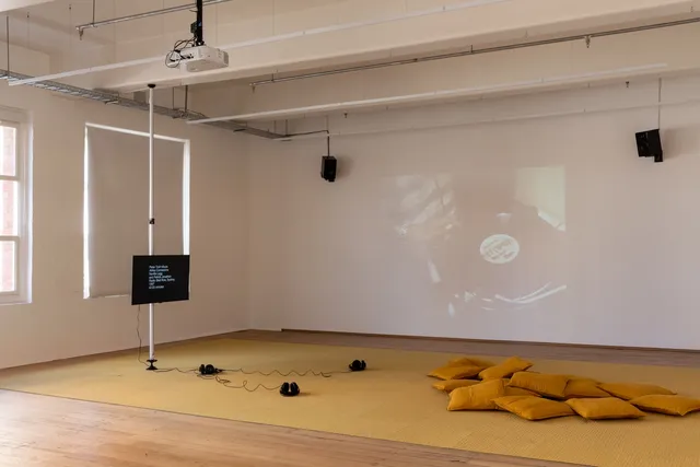 Installation View of 13 Years showing a screen suspended away from the Gallery wall with 3 pairs of headphones and some cushions in front of it. The back wall shows a projection of archival footage of a record sitting on a record player with a person standing over it.