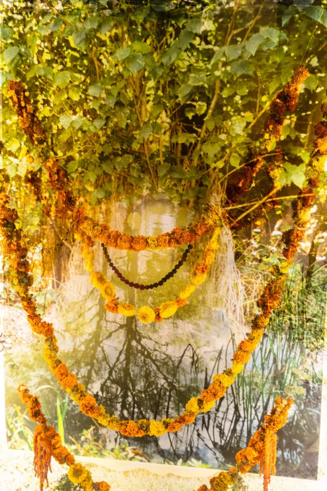 Print of green leaves, trees and water. Four strings of orange and yellow flowers hung across the frame, one beaded string.