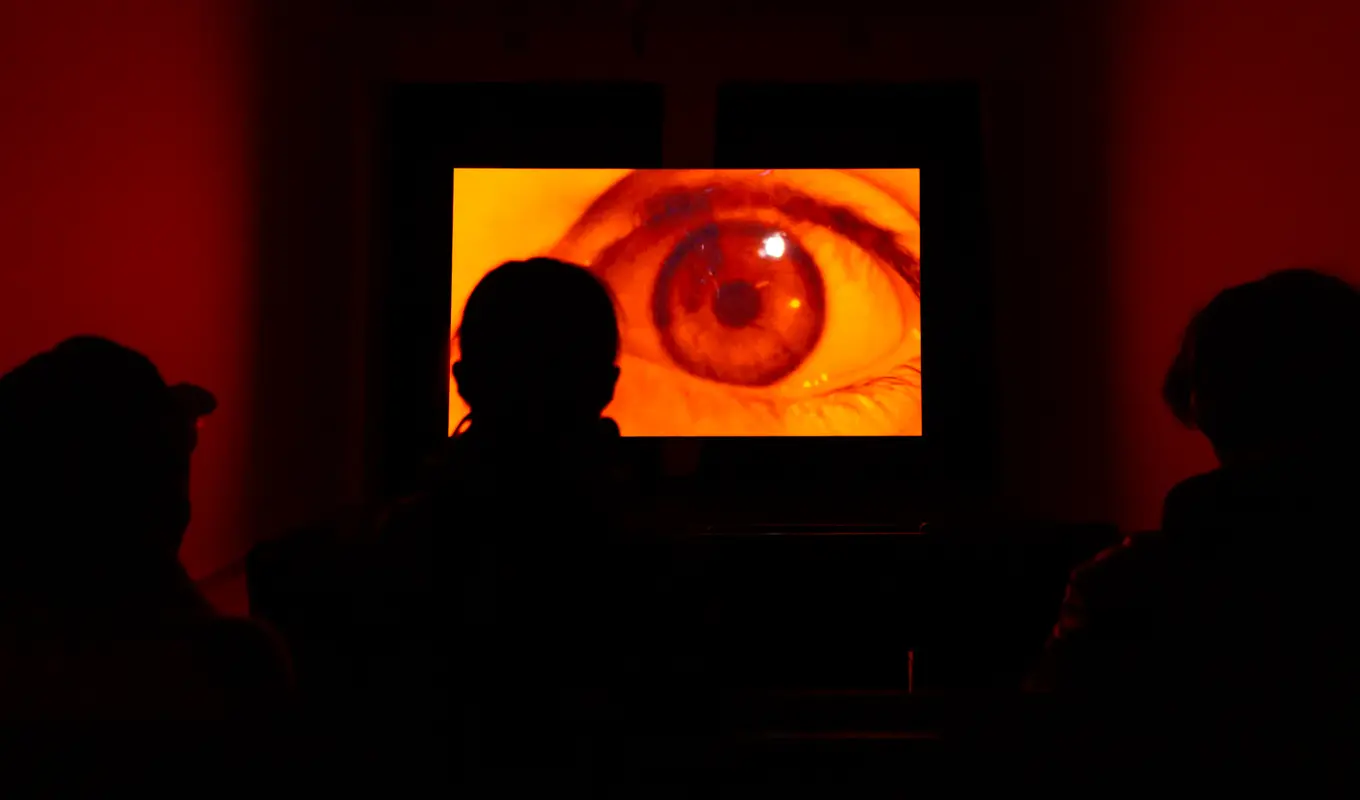 A bright projection of a close up shot of an orange eye illuminates the silhouttes of several figures watching the projection. The eye is widened in shock