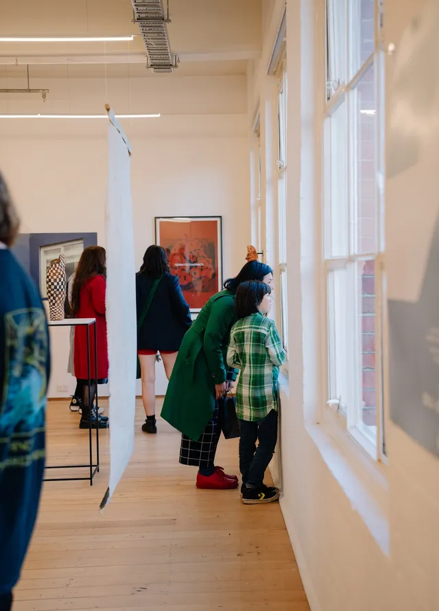 Two figures - a child and an adult - gaze outside a window of the gallery. The works on the distanced wall are undistinguishable.