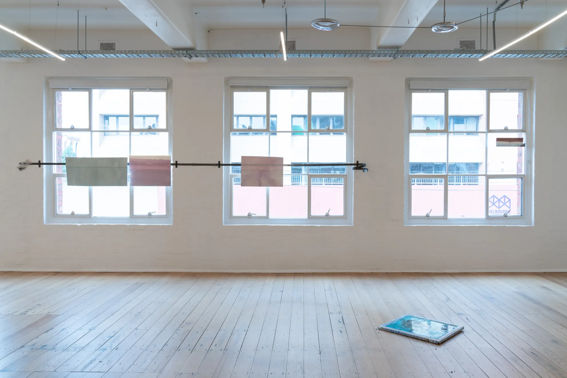 Gallery space, three large windows displaying four paper prints on metal bar. One rectangle blue ceramic work displayed on wood floor.