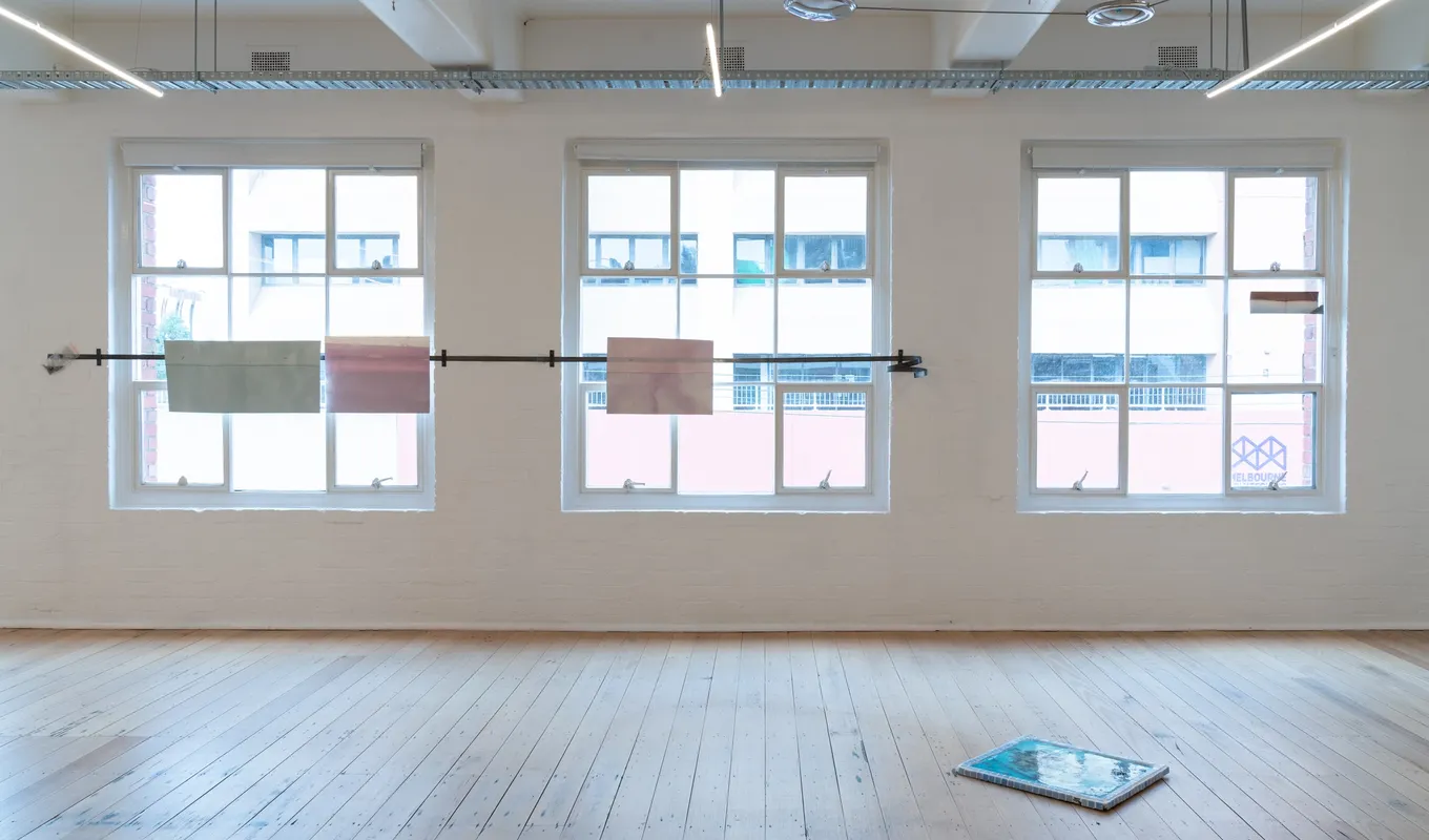 Gallery space, three large windows displaying four paper prints on metal bar. One rectangle blue ceramic work displayed on wood floor.