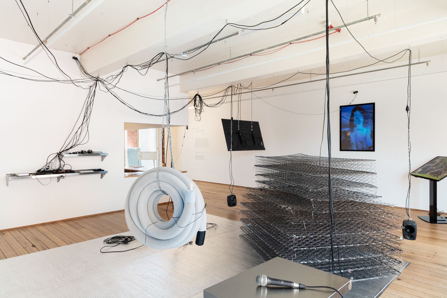 A suspended coil of pipes, a pile of springs from mattresses, two speakers, two screens, an blue image of a woman in the gallery. The middle of the floor is covered in foil mat.