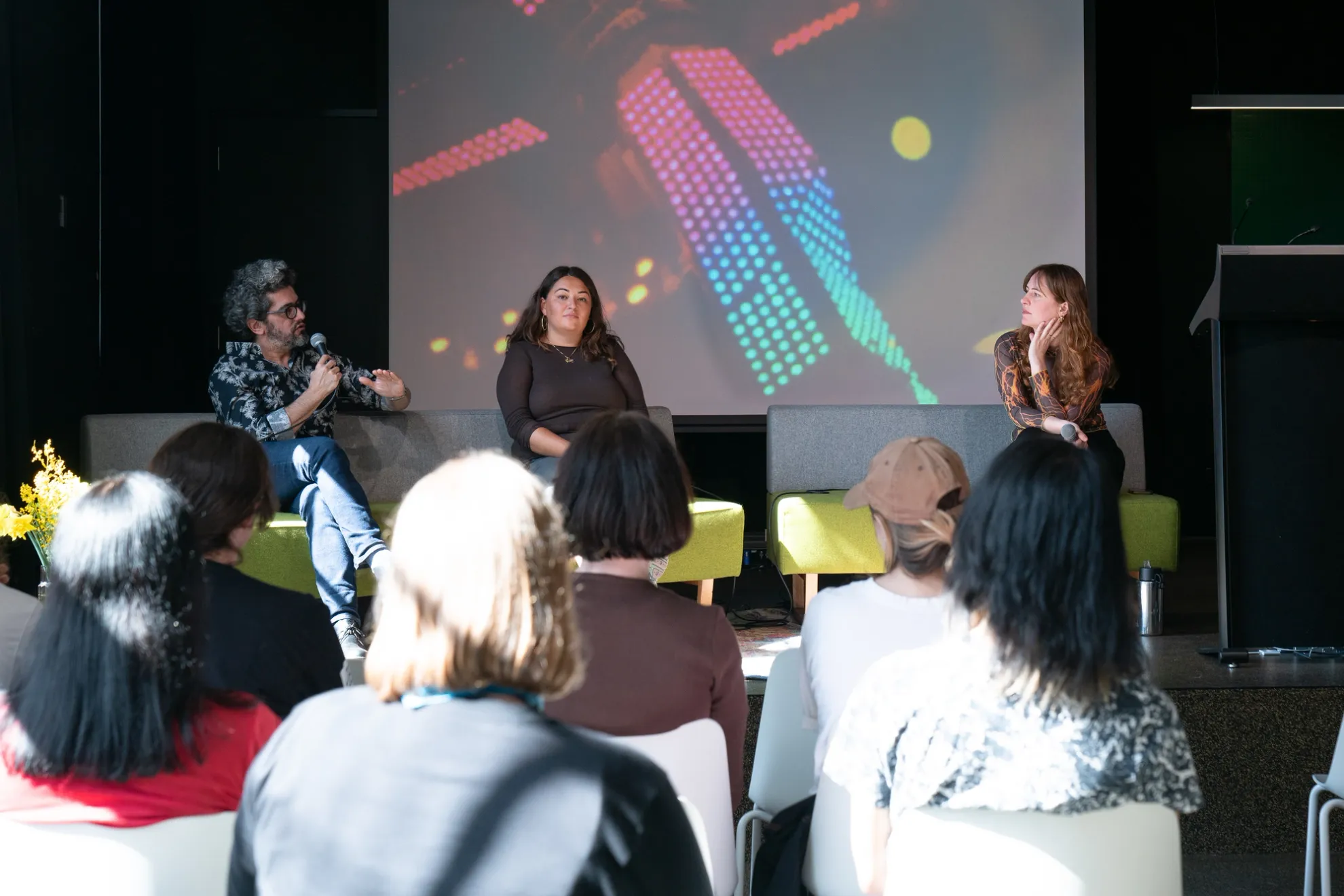 Three people on stage sitting on a couch with a projected image behind them. Man on the left of the couch is speaking to a seated crowd using a microphone.