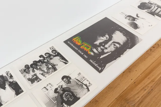 The image shows Archival photos from the collection of Patrick Jonathan sitting on a white shelf. The photos are of the Peter Tosh Australian Tour (1983) taken by Vijay Megan. The photos are black and white and include portraits and groups, additionally there is a poster advertising the tour of Peter Tosh.