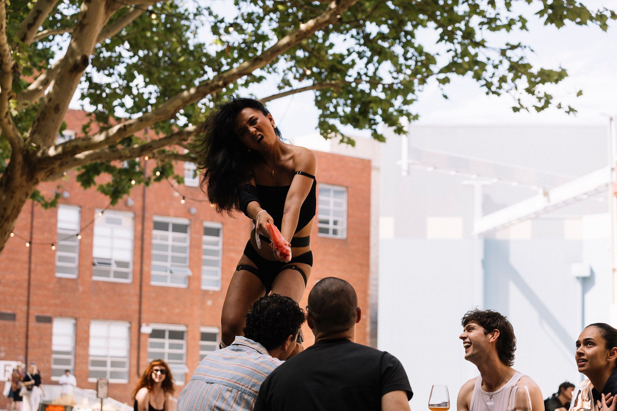 Ari standing on a table holding an orange fish like a gun pointed at someone whose facial expressions are not visible.