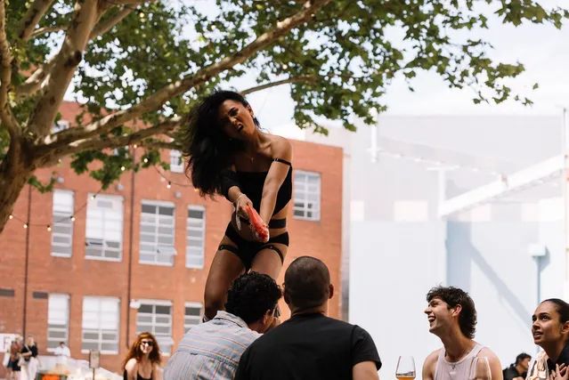 Ari standing on a table holding an orange fish like a gun pointed at someone whose facial expressions are not visible.