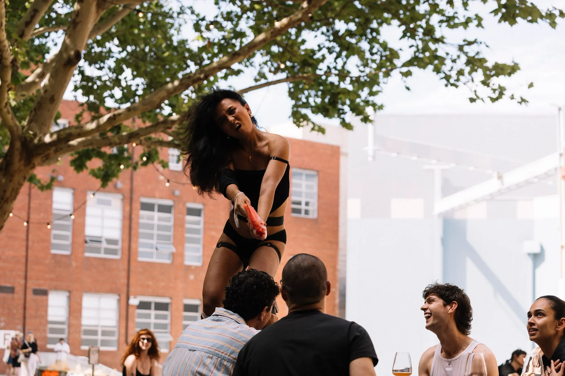 Ari standing on a table holding an orange fish like a gun pointed at someone whose facial expressions are not visible.