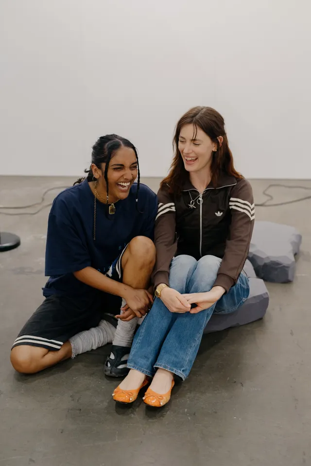 Aarti Jadu and Claire de Carteret sitting on their artwork laughing.