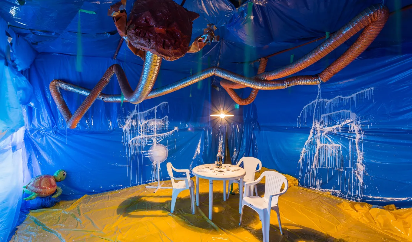A small room-scape - the walls are sheets of vibrant blue plastic, and floor is a yellow plastic sheet and glossy texture. Inside the room is a plastic dining set up - 3 white chairs and a plastic round table. Above the desk, there is a large slinky-like pendant travelling around the ceiling. At the end of the pendant there is a large red bulb, heart-like. On two of the blue plastic walls, there are vandlized, dripping white stains. There is an inflatable turtle in the far corner.