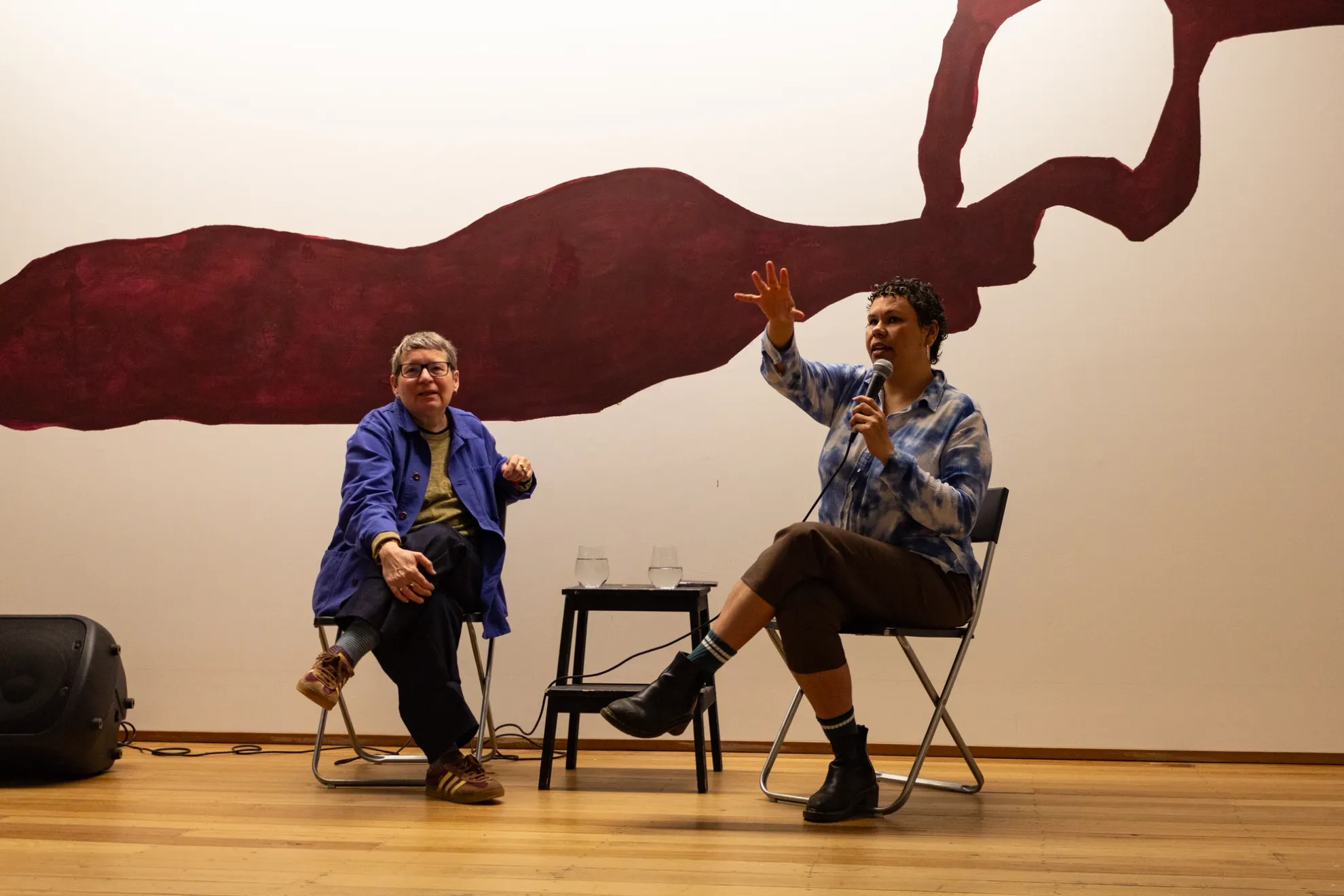 Two individuals sit on fold-out chairs sitting crossed-legged. One is talking into a microphone and has a hand out gesturing. There is a step stood in between them with two glasses of water. A speaker sits to the far left of one of the individuals. A large maroon wall mural can be seen behind them.