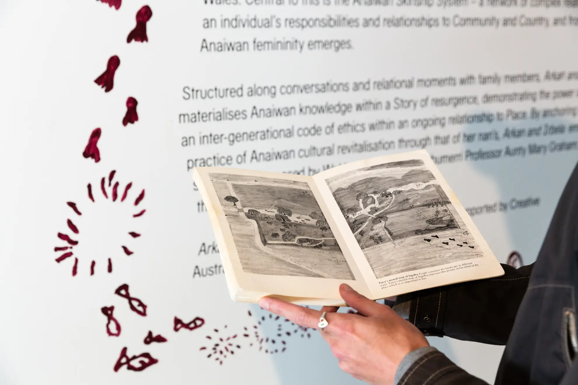 A person holding a book open in front of exhibition text on the gallery wall, small maroon painted drawings surrounding the wall text.