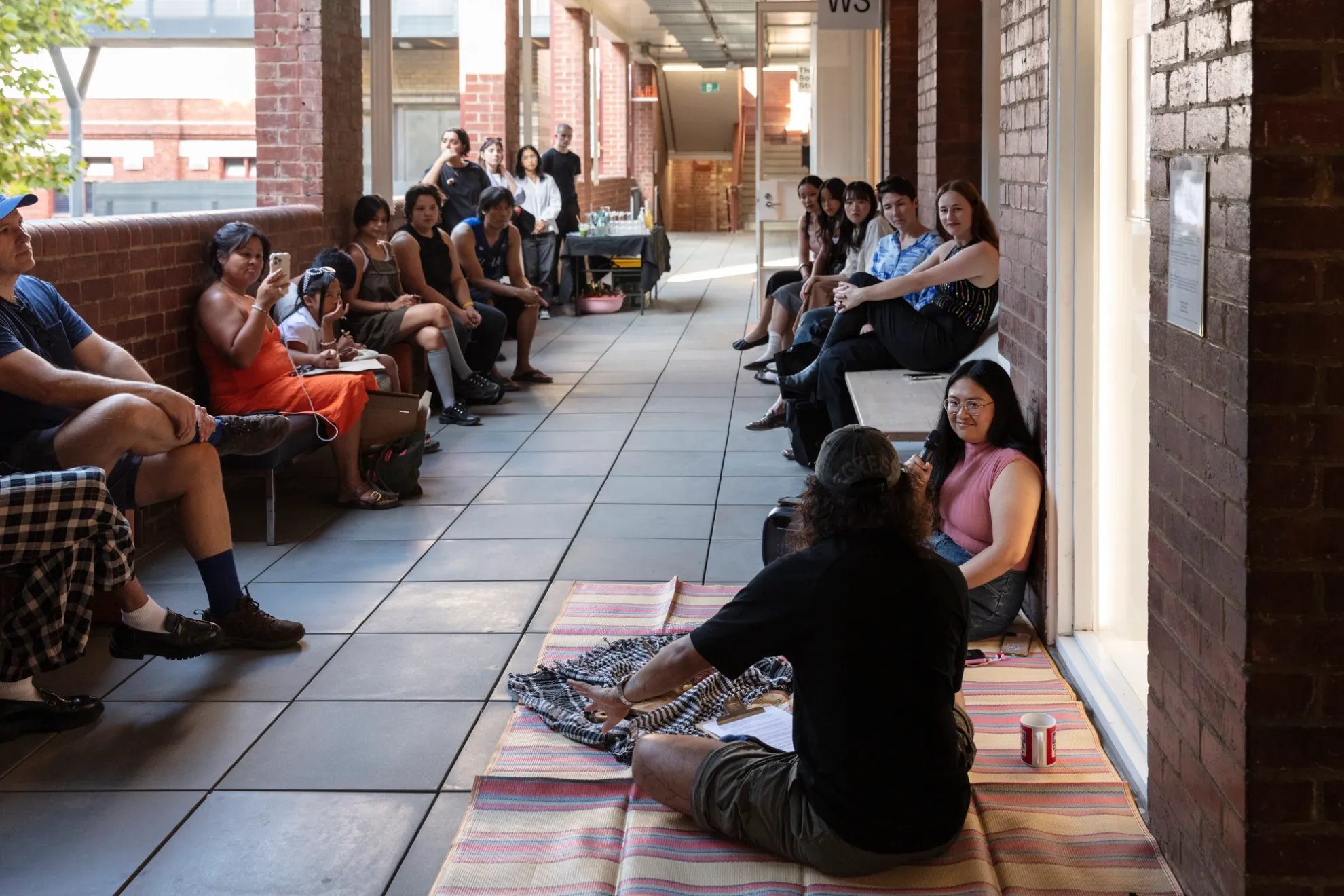 two rows of people sit at the edges of an open corridor, on both sides. There is a carpet in the foreground where tow people are talking. Everyone's gases are directed toward these figures in discussion, who in front of them lies a blanket-like cloth full of plant matter or materials.