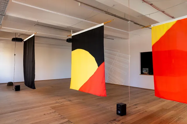 Installation view of "Big Pharmakon". The image shows 3 sheets hanging in the foreground. The left most sheet is completely black while the middle sheet shows a segment of a yellow circle bisected by a red triangle in the bottom right over a black background. The right most sheet is only partially visible and shows a yellow triangle in the top left over a red background. Two speakers sit on the floor between the sheets and a small analog television is visible in the background on a countertop.