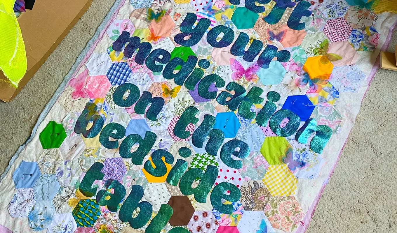 A colourful hexagonal patchwork quilt laying on a carpeted floor. 'I left your medication on the bedside table' is stitched with more fabric onto the quilt.