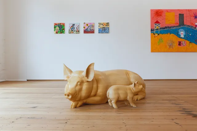 An Installation shot of 'Slime and Ashes' within the West Space Gallery. In the foreground, a Larger mother pig sits next to a little baby pig sculpted with light coloured wood and a semi-gloss finish. The floorboards are a warm wood colour. In the background on the white wall is 4 small, colourful, square paintings with assorted imagery. To the right is a diptych painting, where only one full panel is visible, depicting a city scape meeting a sandy beach. There are characters in motion, the sand is yellow/orange and the sky is pink.
