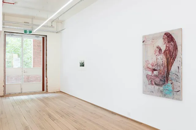 A view of the West Space gallery wall with a small black and white image on the left and a larger painting on the right, depicting two red figures in rough brushstrokes. One figure sits at a table while the other looms behind them.