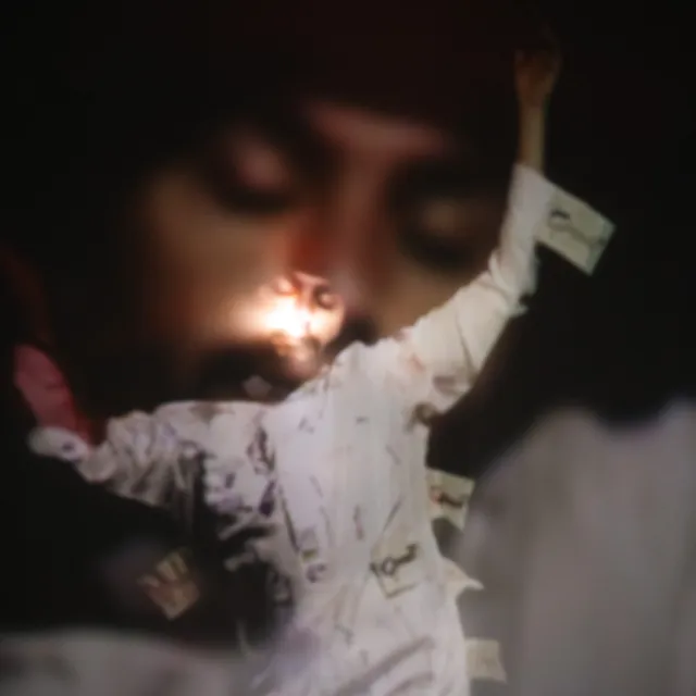 A figure dressed in a white long shirt has their arms raised and their eyes closed. They seem to be mid twirl in a trance. Light is shining on their face.