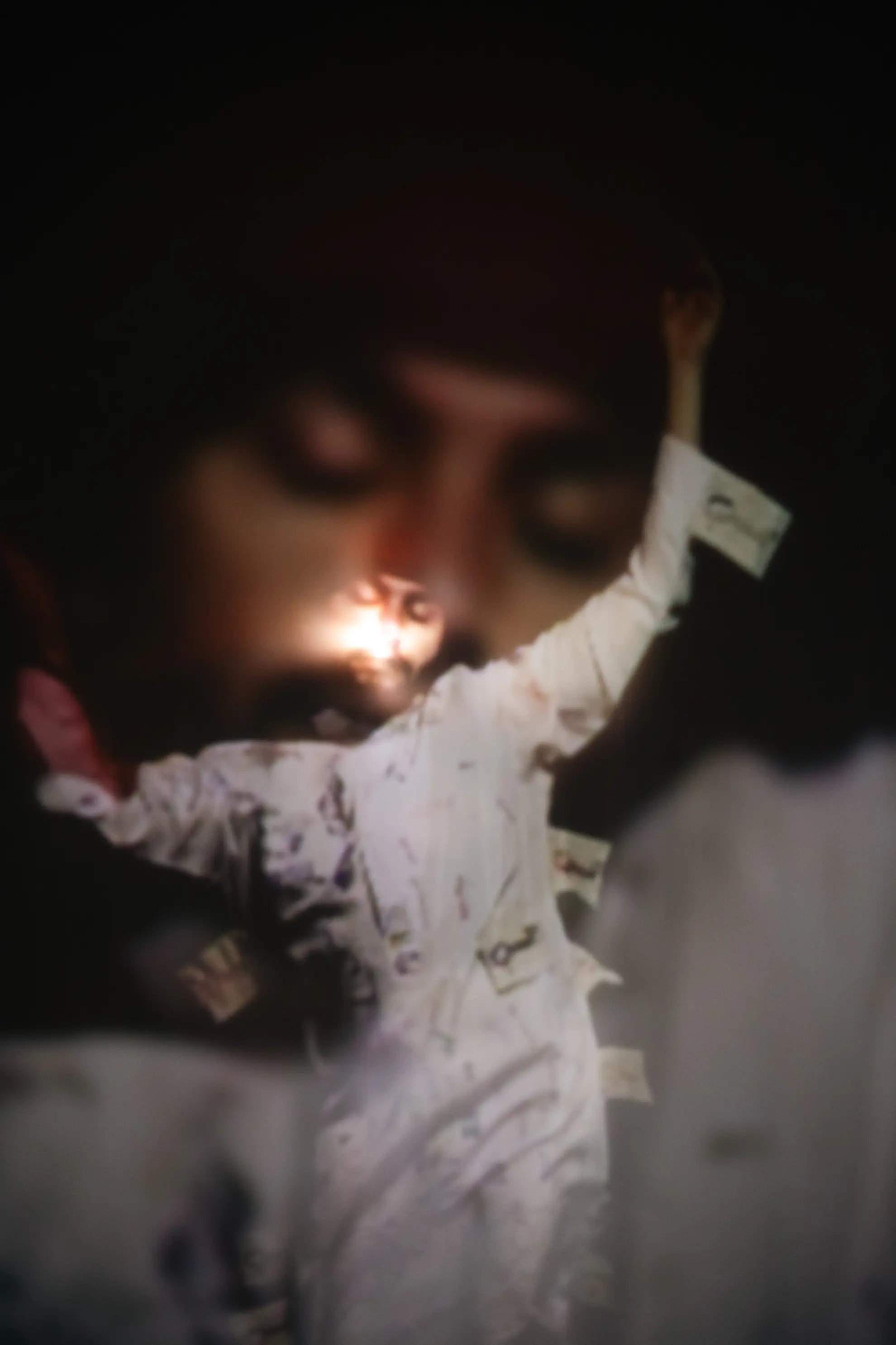 A figure dressed in a white long shirt has their arms raised and their eyes closed. They seem to be mid twirl in a trance. Light is shining on their face.