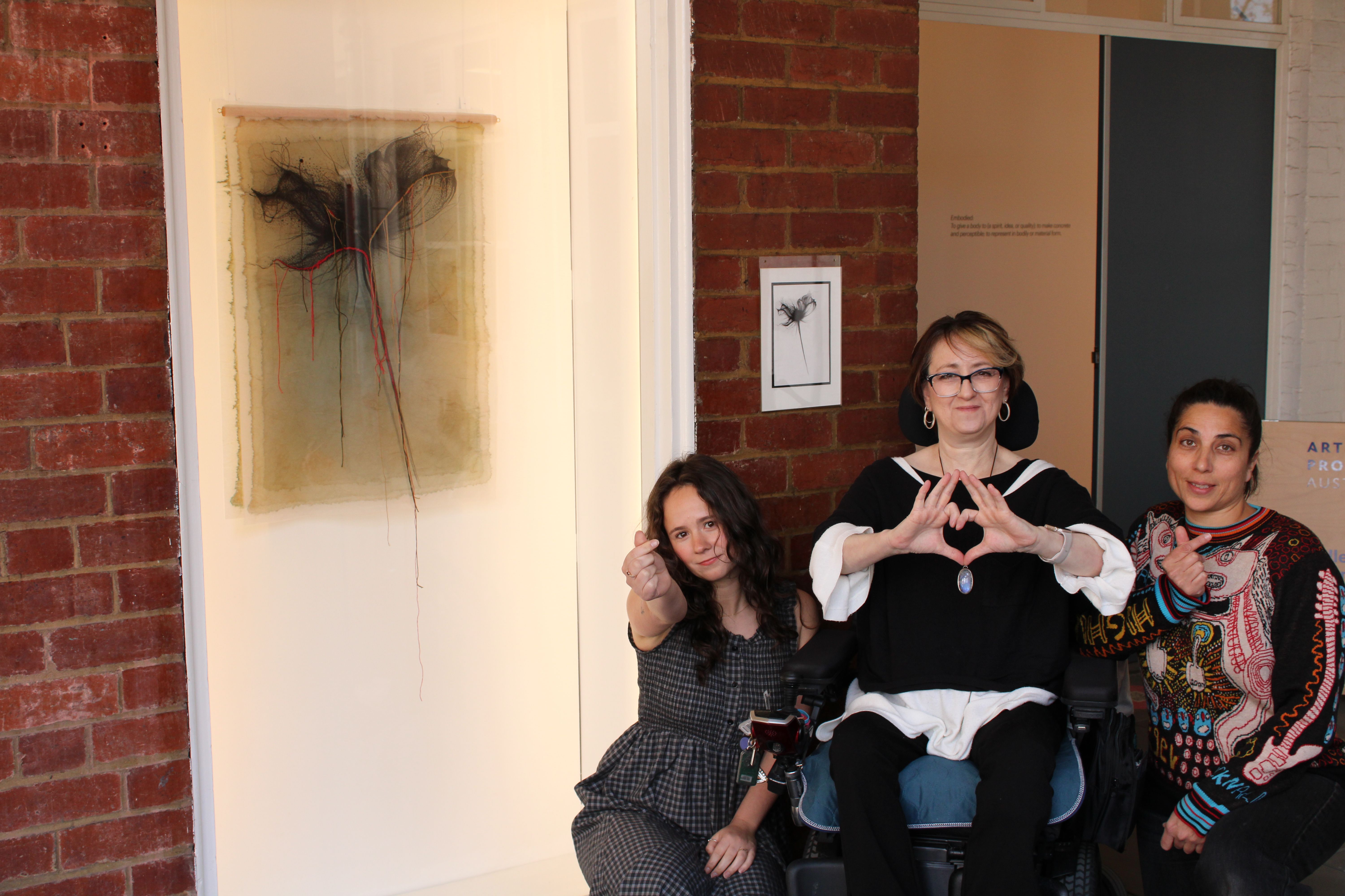 3 people stand in front of the West Space window posing for the camera. The artwork is a textile piece.