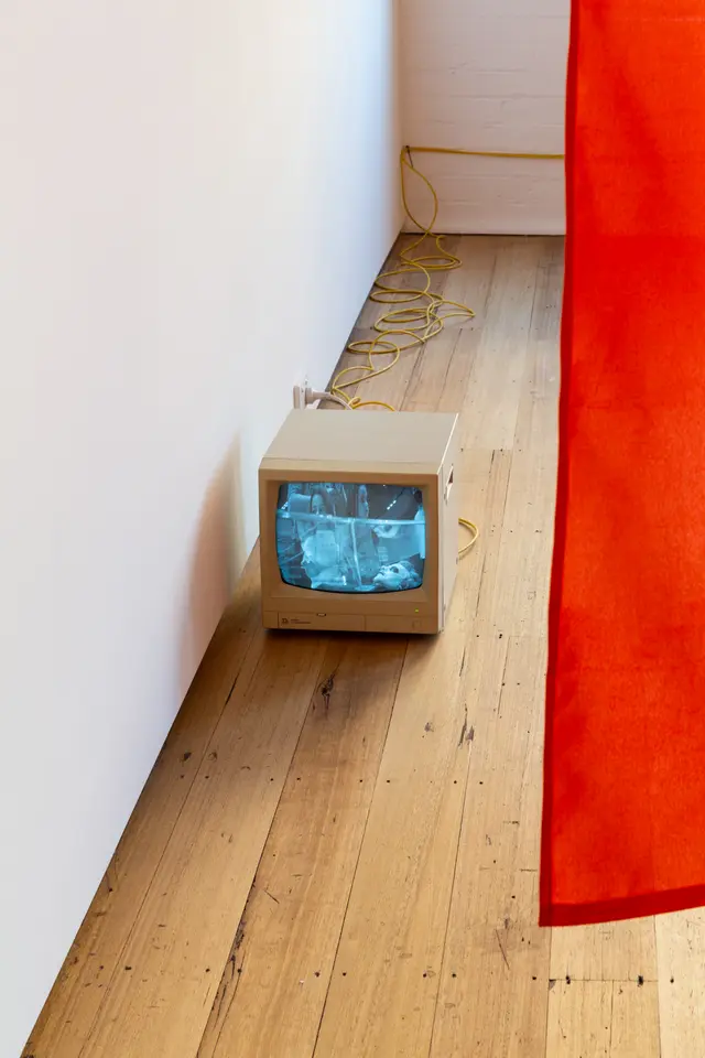 The image shows a small analog television sitting on the ground with a red sheet partially visible on the right side of the image. The screen shows a distorted image of 3 people and is connected to the wall with a bright yellow wire.