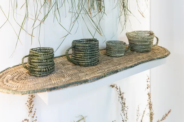 Some densely woven mugs in colours of green, tinted yellow and brown, natural fibres on a rounded, weaved platform. There are 4 mugs. Hung above the elevated mugs is fragments, is the fibre in it's natural state - draping.