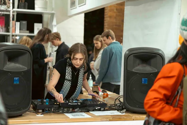 A DJ at West Space is playing music in a crowded space. They are wearing a Palestinian Keffiyeh