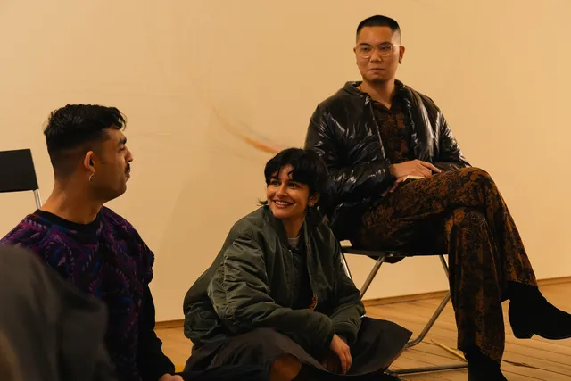 Three individuals are on a stage, discussing with one another. One sits crossed-legged on a chair, and another sits crossed legged on the stage next to him, both with their heads turned to talk to the third person.