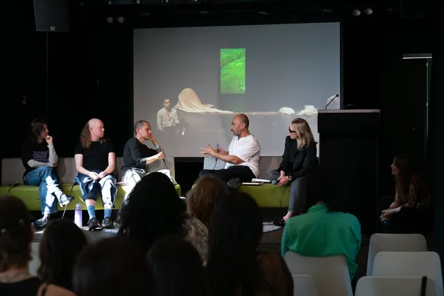 Five individuals are sitting on a green couch on a stage talking with one another. A projection screen is visible behind them. There is a crowd of people seated on white chairs listening.