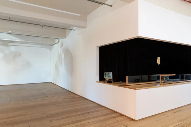 Installation view of "Big Pharmakon". The image shows a countertop with a small analog television and a structure made of wood and glass. A large black curtain hangs behind the counter. To the left of the counter are two projections of a close-up of a person with black and white facepaint.