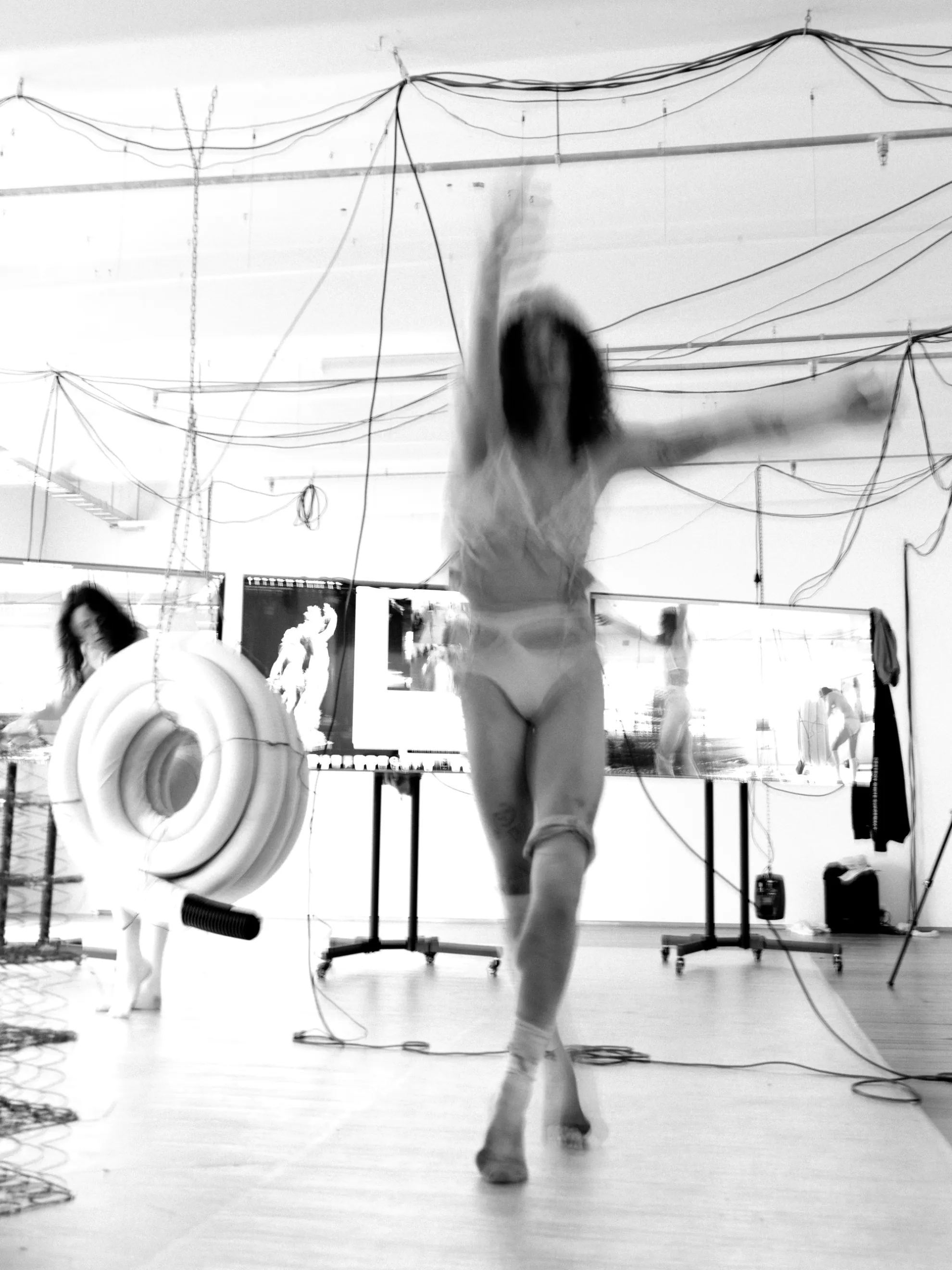 A blurred in motion photograph of Andrea Illés dancing in front of a row of tv screens and wires.