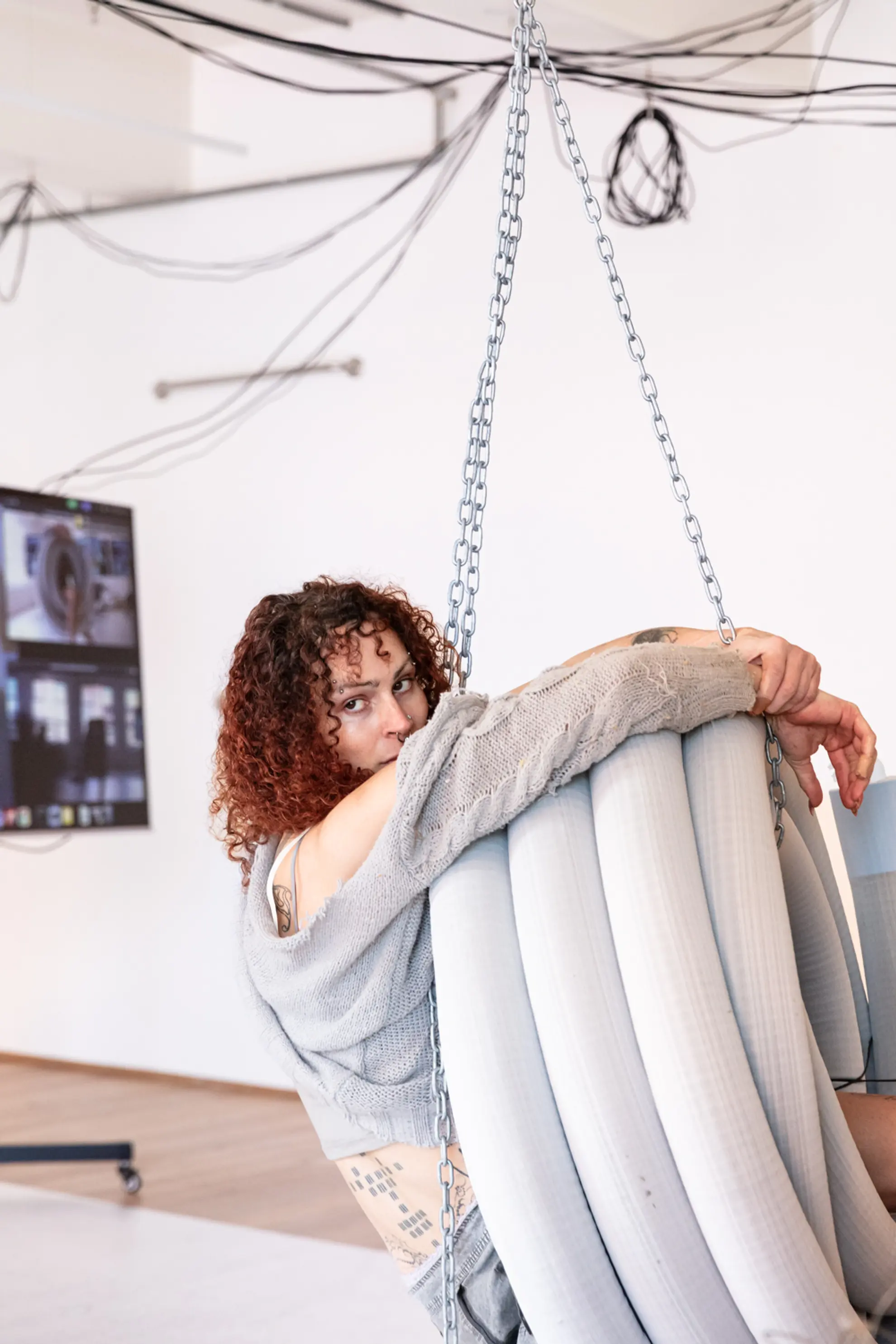 A woman is suspended from the roof hanging in a coiled pipe. She is sitting in the pipe, her arms are holding the pipe, and she is looking at the camera.