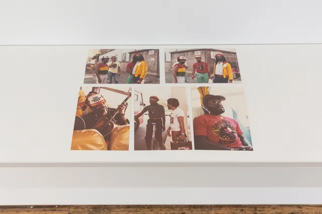 The image shows 5 colour photos sitting on a white shelf. The photos are of the Peter Tosh Australian Tour (1983) taken by Vijay Megan. 2 Photos show a group of 4 men gathered on a street corner and the other 3 photos show people in and around a bus.