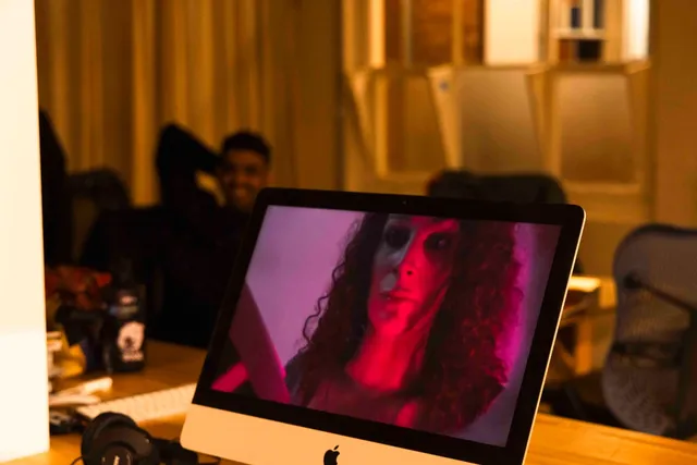A view of Andrea Illes on livestream on a mac computer in the office of West Space Gallery. A blurred figure sits in the background