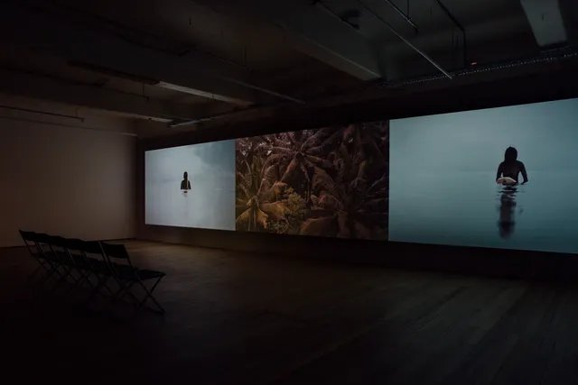 Installation view of a three channel video in the West Space gallery with a row of six chairs facing the work. The first channel shows a female surfer wearing a black wetsuit sitting on her board. The sky and ocean are a pale grey with clouds gathering in the top right corner. The second channel depicts palm trees and the third shows another female surfer on her board with a darkening blue sky and ocean.