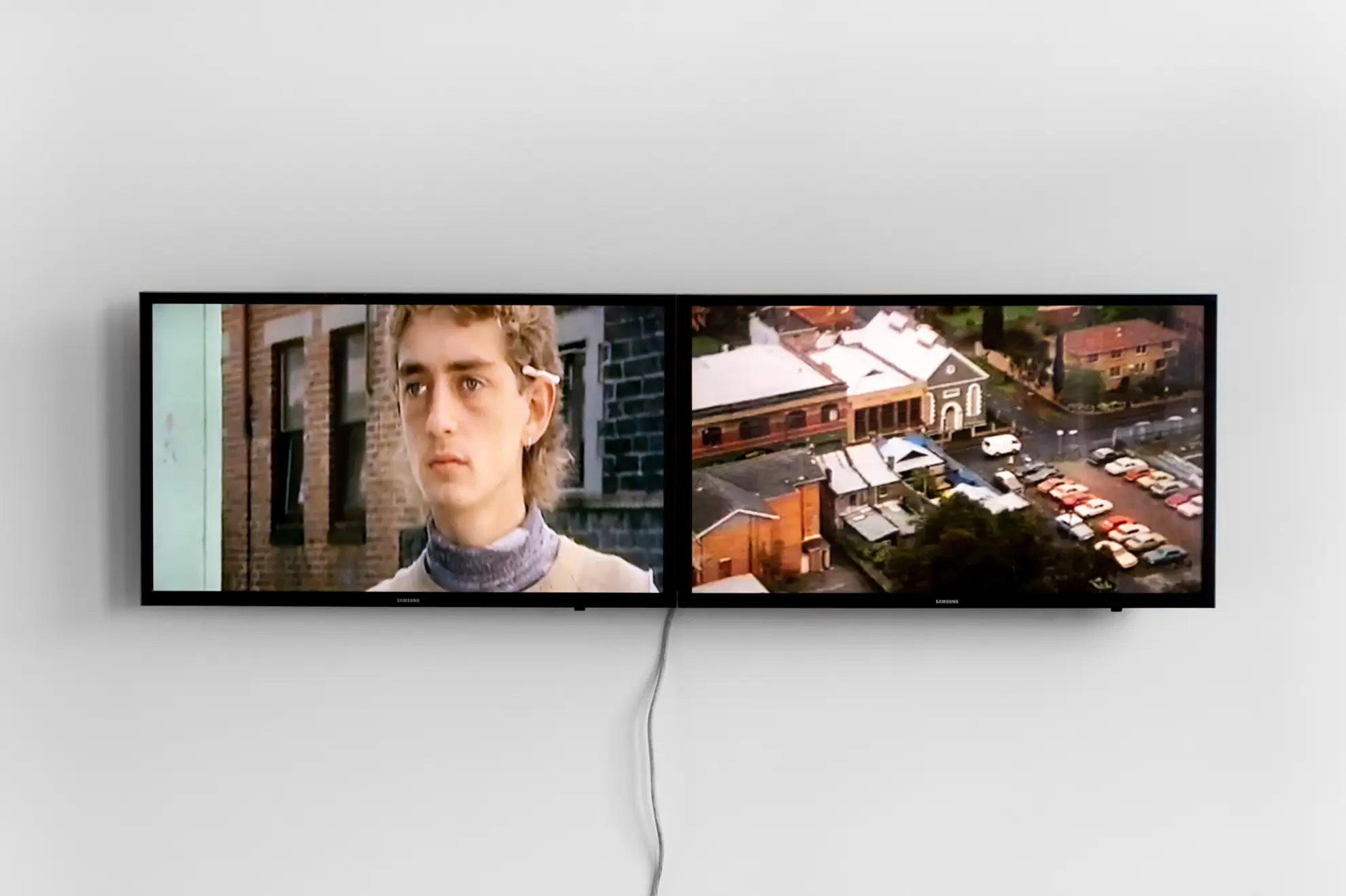 Two rectangular screens sit side by side, hanging on a white wall. The left screen shows a person looking off to the side, with blonde curly hair and a cigarette behind their ear. On the right screen is an aerial view of several buildings and a busy carpark.