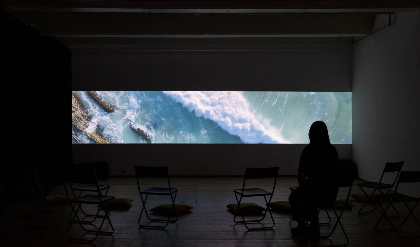 A shadowed figure sits in front of a thin format projection the length of a large wall depicting a heavy swell of an ocean