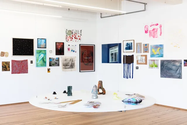 Two large walls of the west space gallery are installed full of artworks. In the foreground, a large circular white suspended shelf hangs in space adorned with many small sculptural artworks.