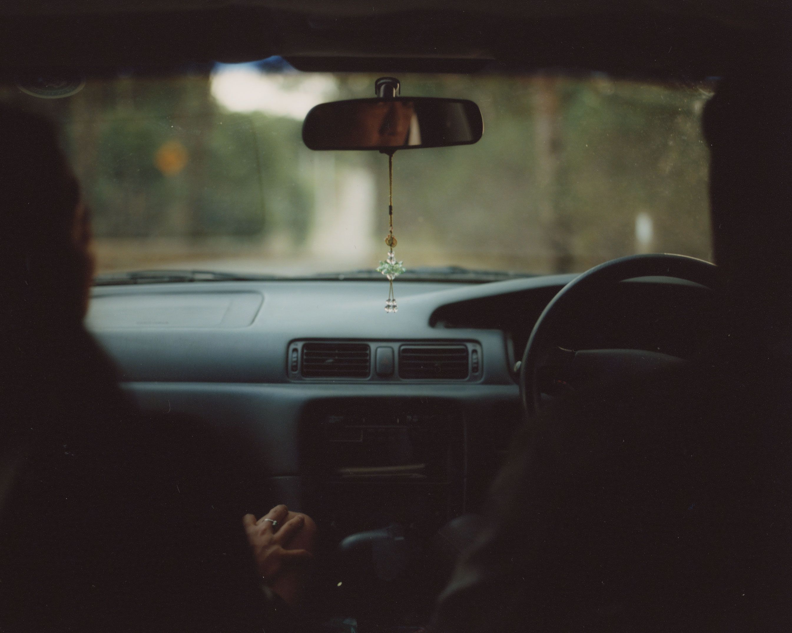 View through the front two seats of a car with two people sitting in them. A blurry view of trees lies beyond the car window.