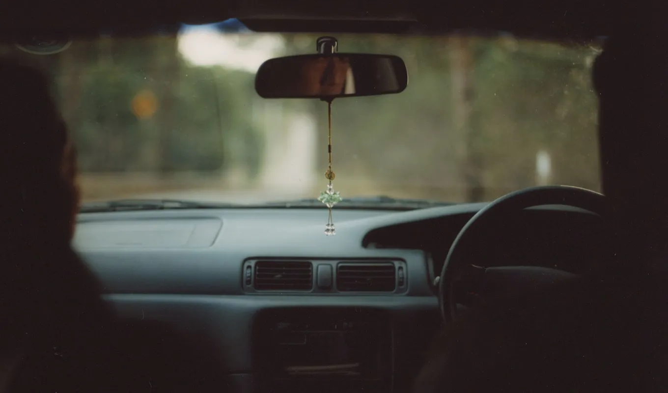 View through the front two seats of a car with two people sitting in them. A blurry view of trees lies beyond the car window.