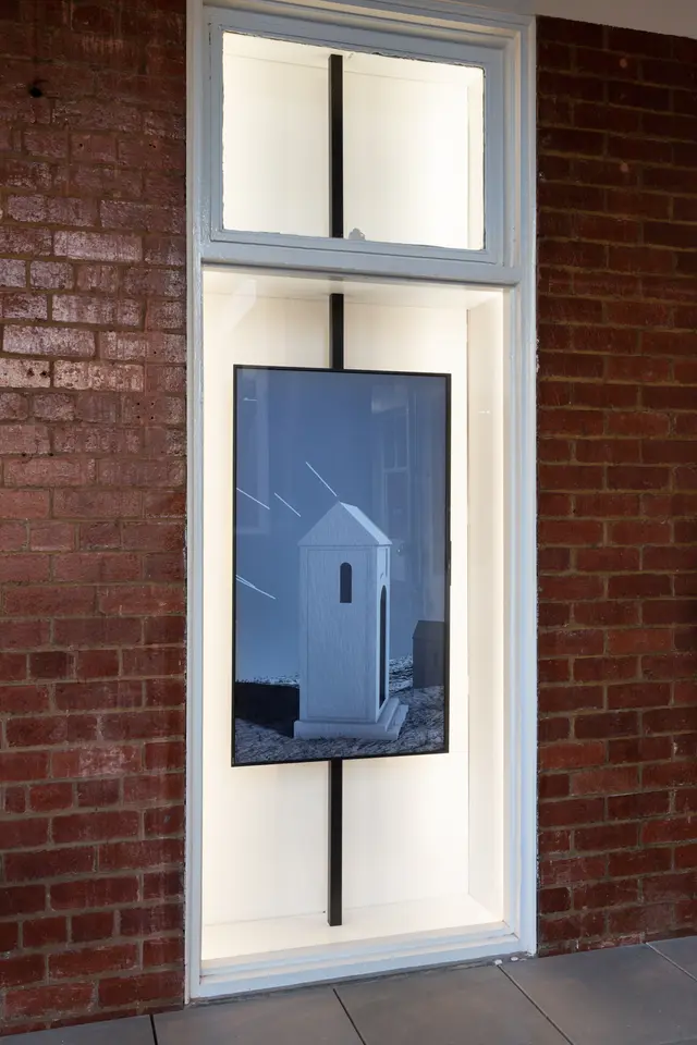 ID: A rectangular screen suspended on a pole inside a glass vitrine: the West Space Window. The screen depicts a grey animated image of a chapel-like structure with one window off to the side just underneath its roof. Another similar structure sits in the background along with four lines suspended in the air.