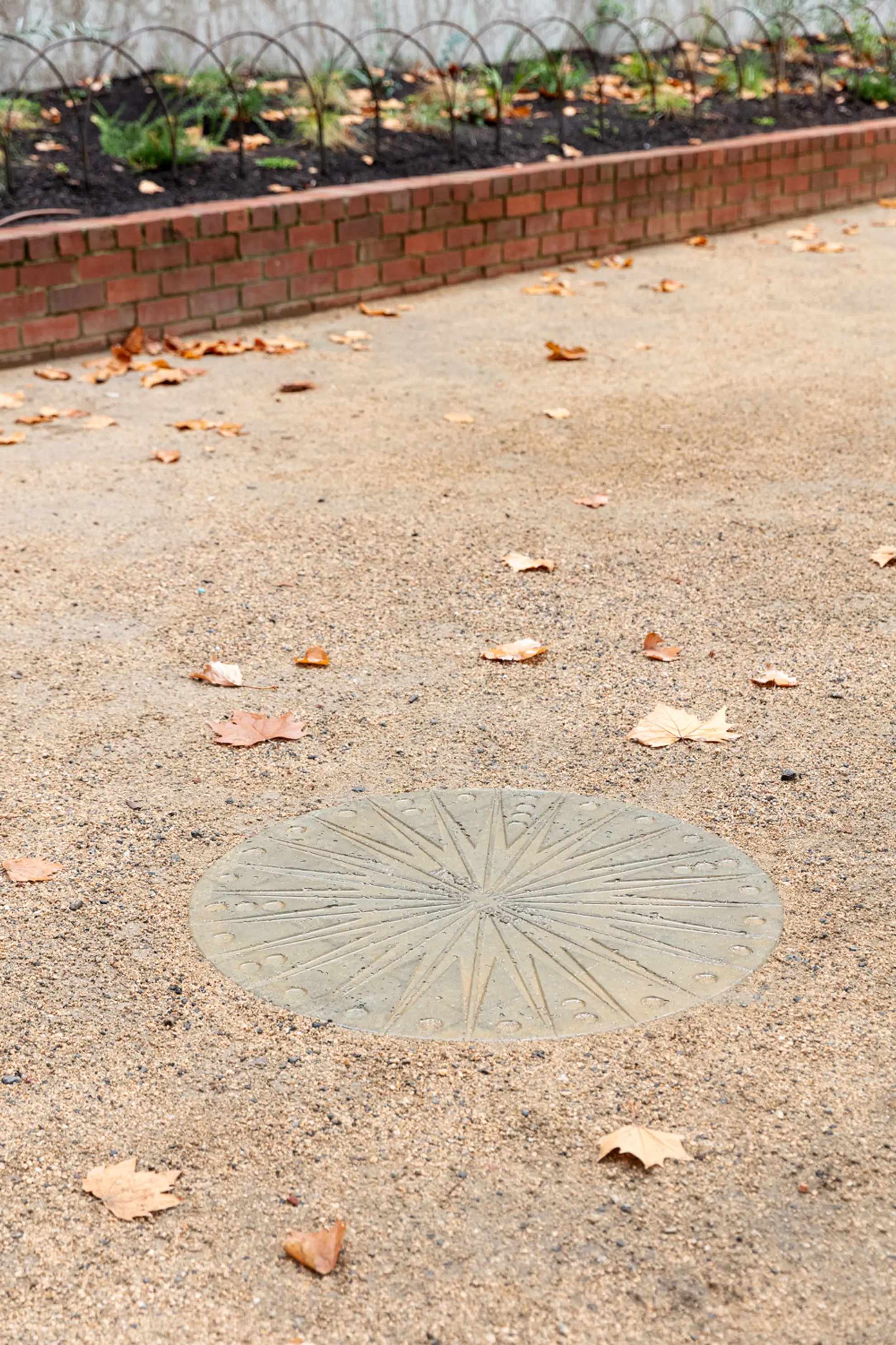 A circular concrete slab engraved with multiple star points nestled into an outdoor gravel floor. Autumn leaves are scattered around it and in the background a short brick wall and some green plants can be seen.