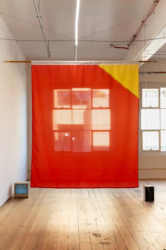 Installation view of "Big Pharmakon". The image shows a semi-transparent sheet in front of a window in West Space Gallery. The sheet is red with a yellow triangle in the top right corner. A small analog television and a speaker sit on either side of the sheet on the floor.