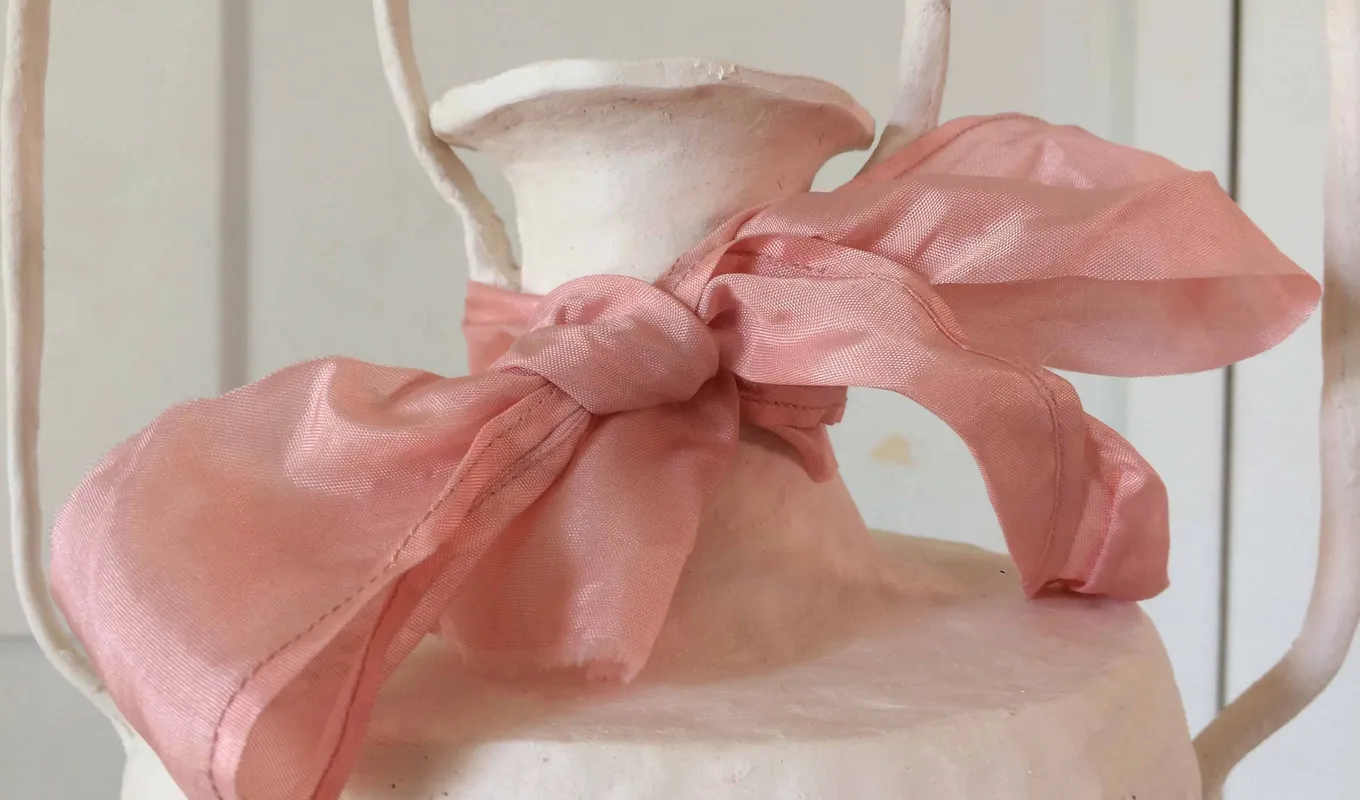 Close-up of a large pink fabric bow tied around the neck of a white textured vase.